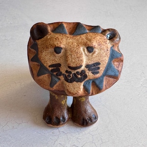 May include: A ceramic lion figurine with a textured, speckled brown body and a detailed face. The lion has black accents on its mane, eyes, and mouth, and stands on two small feet. The figurine is a decorative item.