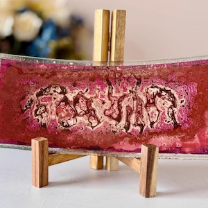 May include: A rectangular glass decorative tray with a curved shape, featuring a maroon and pink abstract design. The tray is displayed on a small wooden stand. The background includes blurred flowers.