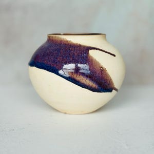May include: A small, round ceramic vase with a wide, open mouth. The base of the vase is a matte, off-white cream color. A glossy, deep purple and dark blue glaze sweeps across the upper portion of the vessel in a diagonal, organic pattern.