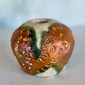 May include: A small, round ceramic vase with a wide opening. The surface features a textured, metallic copper and bronze glaze with iridescent highlights. Deep green, crackled glaze accents run through the center, contrasting with the metallic finish.