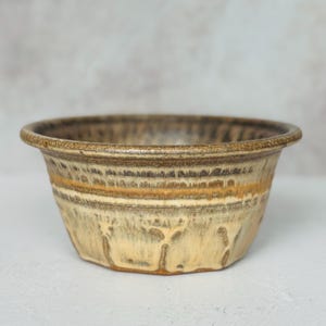 May include: A ceramic bowl featuring a wide, rounded rim and a tapered base. The exterior displays a reactive glaze with layered tones of cream, beige, and dark brown, creating a textured, dripped appearance that transitions from dark at the top.