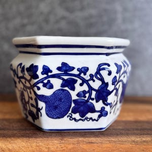 May include: White and blue ceramic planter with a hexagonal shape. The planter features a detailed blue floral and fruit design. The rim is white with a blue stripe. The planter sits on a wooden surface.