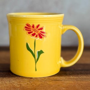 May include: A bright yellow ceramic mug featuring a single stylized flower graphic on the front. The flower has red petals with a dark center and a green stem with leaves. The mug has a smooth, glossy finish, a rounded handle, and a ribbed rim.