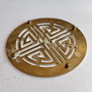 May include: A round, brass-colored trivet with a geometric, openwork design. The trivet has a series of small, raised feet and a label that reads "Made in Hong Kong". The design is a repeating pattern of interlocking shapes.