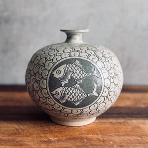 May include: A round ceramic vase with a narrow neck, featuring a dark gray circular medallion on the front containing two white fish figures. The rest of the vase is decorated with a repeating pattern of small, swirling, cloud-like white lines.
