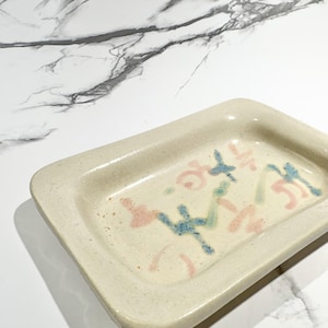 May include: A rectangular ceramic dish with rounded corners. The dish is off-white with a speckled texture. It features abstract designs in pink, blue, and green. The dish is likely handmade and suitable for serving small items.