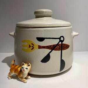 May include: White ceramic crock with a lid, featuring a brown, yellow, and black design of kitchen utensils. A small orange and white ceramic cat figurine is in the foreground.