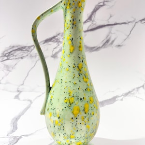 May include: A tall, light green ceramic pitcher with a slender neck and a curved handle. The pitcher is decorated with yellow and black speckles. It has a round base and a spout for pouring. The pitcher is likely used for serving beverages.
