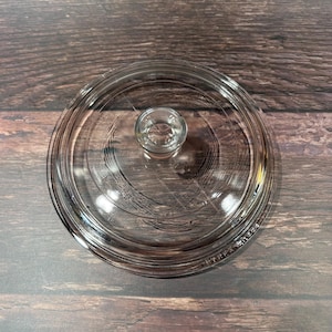 Könnte beinhalten: Ein klarer Glasdeckel mit einem kleinen, runden Griff. Der Deckel ist kreisförmig mit einer leicht gewölbten Form und ist für einen Topf oder eine Pfanne konzipiert. Das Glas ist transparent.