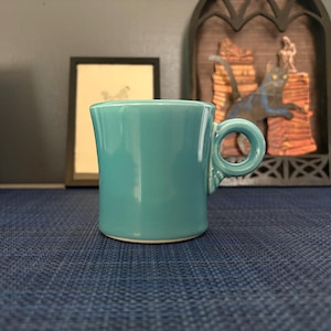 May include: A turquoise ceramic mug with a rounded handle. The mug has a smooth, glossy finish and a simple, classic design. The mug is approximately 3 inches tall.