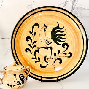 Puede incluir: Un plato grande y redondo de cerámica con un diseño de gallo. El plato tiene un fondo beige con detalles en negro y un borde negro. También se ve una pequeña jarra de cerámica a juego con tapa y diseño de gallo.
