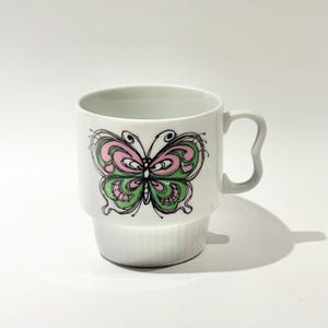 Puede incluir: Taza de cerámica blanca con un diseño caprichoso de mariposa en rosa y verde. La mariposa está delineada en negro, agregando contraste al diseño. La taza tiene una base apilada única y un asa curva.