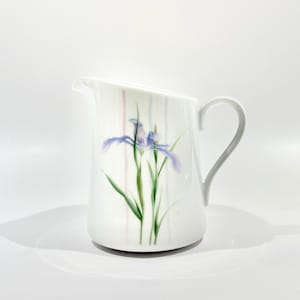 May include: White ceramic pitcher with a curved handle and spout. The pitcher features a painted design of a purple iris flower with green leaves and vertical pastel stripes. The pitcher is approximately 6 inches tall.