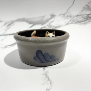 May include: A gray ceramic pot with a dark interior and a blue abstract design. A small, detailed figurine of a fox is inside the pot, with its head and tail visible. The pot has a rustic, handmade appearance.
