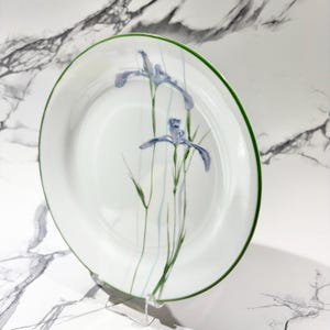 May include: White ceramic plate with a green rim and a watercolor design of blue iris flowers and green stems. The plate is displayed on a clear stand, showcasing the floral artwork.