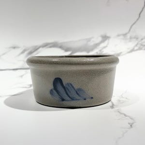 May include: A small, round, gray stoneware bowl with a slightly textured surface. The bowl features a simple blue painted design on the side. The rim is rounded, and the bowl sits on a white surface.