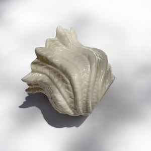 May include: A cream-colored, ceramic seashell sculpture with a textured surface. The shell has a detailed, ridged design and is set against a white background. The sculpture's shadow is visible.