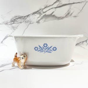 May include: A white rectangular serving bowl with blue floral designs. The bowl has handles on each side and a decorative blue floral pattern. A small ceramic cat figurine is in front of the bowl.