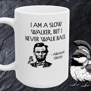 Może przedstawiać: Biały ceramiczny kubek z cytatem "I AM A SLOW WALKER, BUT I NEVER WALK BACK" i portretem Abrahama Lincolna. Czarno-biała ilustracja ptaka w hełmie znajduje się po prawej stronie.