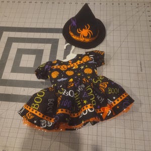 May include: A black dress with short puff sleeves and a flared skirt featuring a Halloween print of orange pumpkins, bats, stars, and the word BOO in various colors. The dress includes an orange ribbon trim and a matching black witch hat.