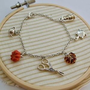 Puede incluir: Un brazalete de plata con varios dijes, incluyendo una tetera, una calabaza, unas tijeras, una hoja, una linterna y una tortuga. Los dijes están unidos a una cadena de plata.