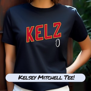 May include: Black t-shirt with the name "KELZ" in red letters with a yellow outline, and the number "0" in white. The text "Kelsey Mitchell Tee!" is displayed in a white banner at the bottom of the shirt.