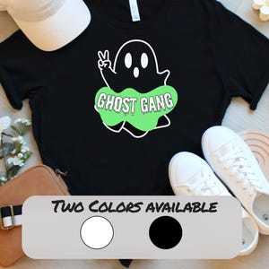 May include: Black t-shirt featuring a white ghost graphic with the text "GHOST GANG" in a green bubble. The shirt also has the text "TWO COLORS AVAILABLE" with a white and black circle. White sneakers are visible.
