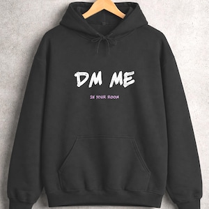 May include: Black hooded sweatshirt with a front pocket and drawstring. The sweatshirt displays the white text "DM ME" above the purple text "IN YOUR ROOM". The hoodie is suspended on a wooden hanger.