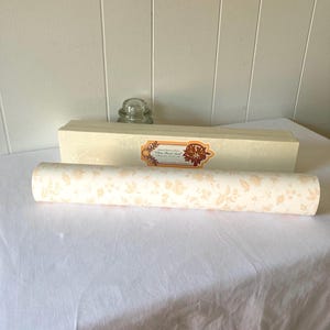 May include: A rolled-up, cream-colored wallpaper with a repeating floral pattern in a soft peach hue. A rectangular box with a decorative label and a glass object are also present.