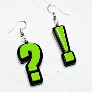 May include: A pair of earrings featuring a green question mark and a green exclamation point. The earrings are black outlined and have silver hooks.