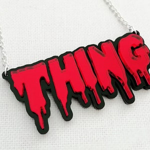 May include: A silver chain necklace with a red and black acrylic pendant that says "THING" in dripping letters.