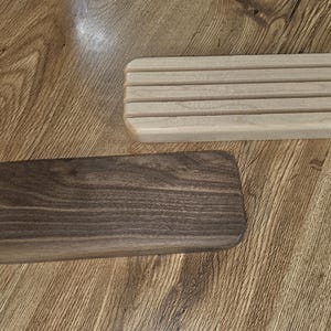 May include: Two wooden soap dishes. One is a light-colored rectangular tray with parallel grooves. The other is a dark brown, solid rectangular tray. Both are on a wooden surface.