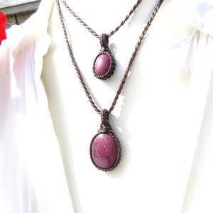 May include: A layered necklace with two pendants featuring a deep pink, oval-shaped gemstone. The necklace is made with brown cord and the pendants are knotted with a macrame style.