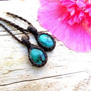 Könnte beinhalten: Zwei türkisfarbene Edelsteinanhänger mit brauner Makramee-Kordel, vor einer verwitterten weißen Holzoberfläche. Im Hintergrund eine leuchtend rosa Blume. Die Anhänger haben ovale türkisfarbene Steine.