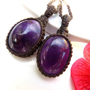 May include: Two purple amethyst gemstone pendants with a brown macrame cord. The pendants are oval shaped and have a shiny, polished finish.