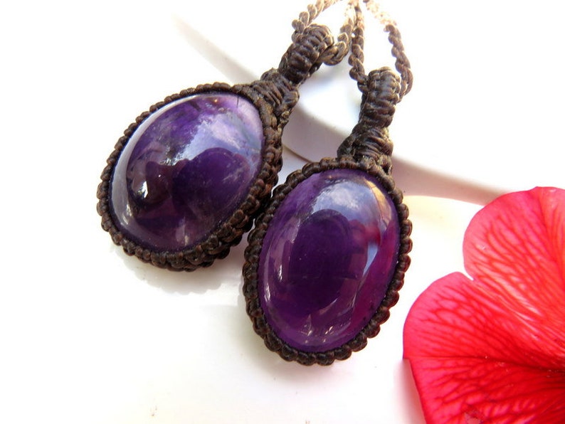 May include: Two purple amethyst gemstone pendants with a brown macrame cord. The pendants are oval shaped and have a shiny, polished finish.