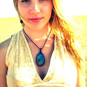 May include: A woman with long blonde hair wearing a light beige sleeveless top with a dark blue oval gemstone pendant necklace.