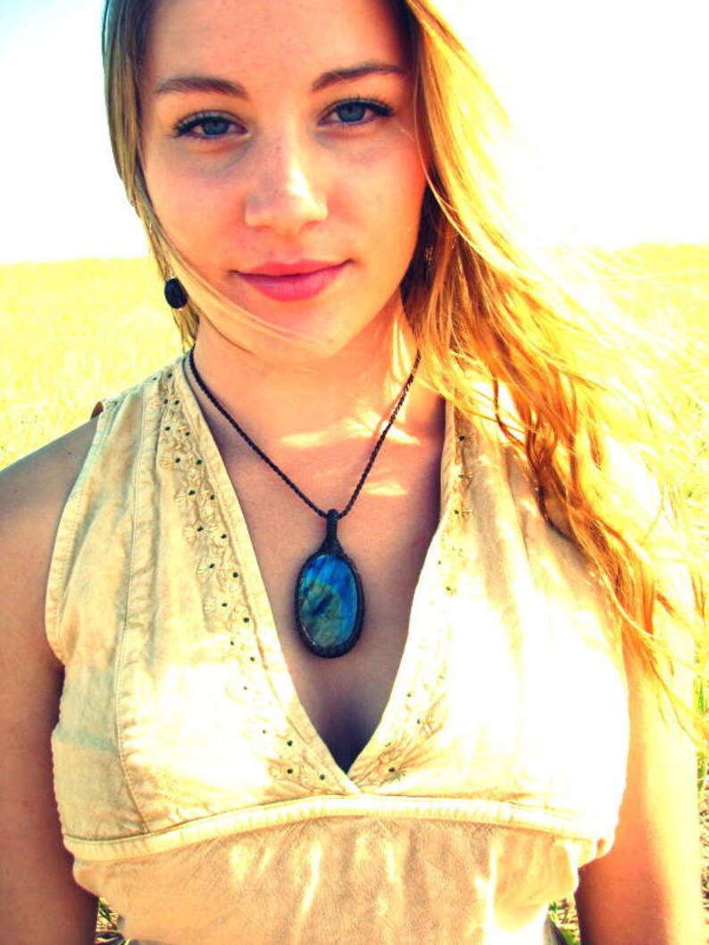 May include: A woman with long blonde hair wearing a light beige sleeveless top with a dark blue oval gemstone pendant necklace.