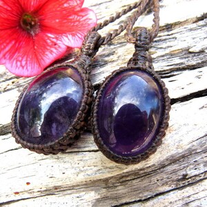 May include: Two purple amethyst gemstone pendants on brown macrame cord necklaces. The pendants are oval shaped and have a shiny, polished finish.