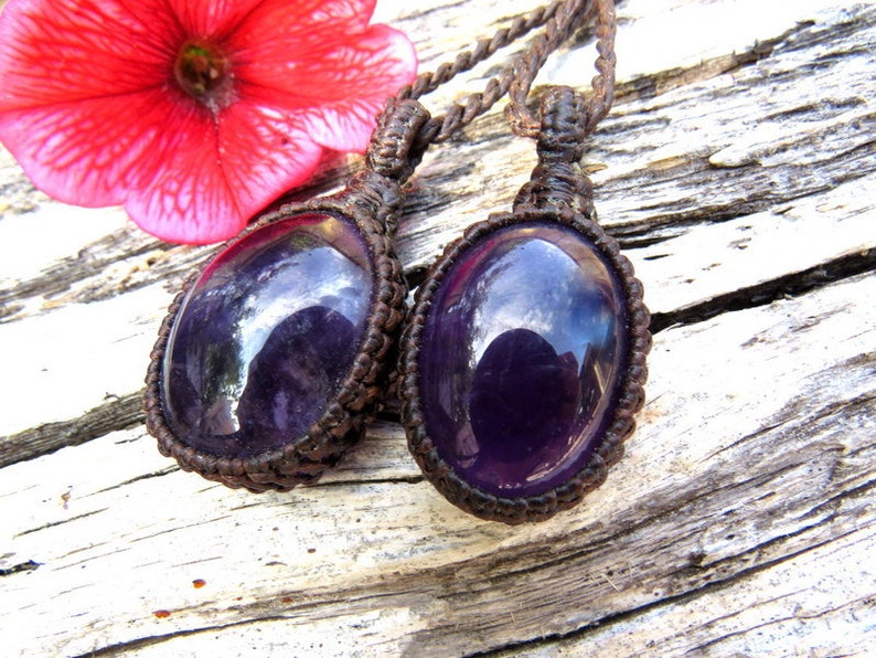 May include: Two purple amethyst gemstone pendants on brown macrame cord necklaces. The pendants are oval shaped and have a shiny, polished finish.