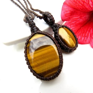 Puede incluir: Dos collares de cordón de cuero marrón con colgantes ovalados de ojo de tigre. El colgante más grande mide aproximadamente 5 cm de largo y el colgante más pequeño mide aproximadamente 2,5 cm de largo.