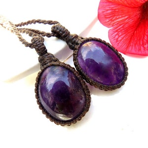 May include: Two purple amethyst stones are tied to a brown cord necklace. The stones are oval shaped and have a shiny, polished finish.
