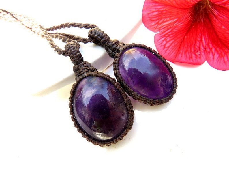 May include: Two purple amethyst stones are tied to a brown cord necklace. The stones are oval shaped and have a shiny, polished finish.