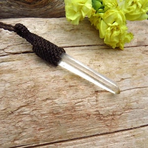 Puede incluir: Un colgante de cristal de cuarzo transparente con un cordón tejido marrón. El cristal tiene forma puntiaguda en un extremo y está envuelto en un material tejido marrón. El fondo es una superficie de madera con flores amarillas.