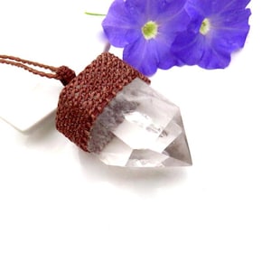 May include: A clear quartz crystal pendant with a brown woven cap and cord. The crystal is faceted and pointed. Two purple flowers are in the background. The pendant is on a white surface.