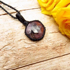 May include: A handmade necklace featuring a faceted, reddish-purple gemstone pendant. The pendant is encased in a black woven setting and hangs from a black cord. The background includes yellow roses on a light-colored wooden surface.