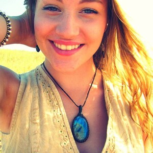 May include: A woman with long blonde hair is wearing a light brown sleeveless top with a blue and green gemstone pendant necklace.