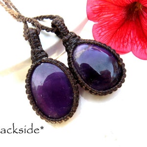 May include: Two purple amethyst stones are wrapped in brown cord and tied together. The stones are oval shaped and have a shiny, polished finish. The cords are knotted and tied together at the top, creating a pendant.