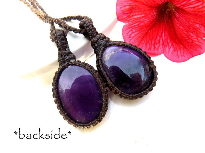 May include: Two purple amethyst stones are wrapped in brown cord and tied together. The stones are oval shaped and have a shiny, polished finish. The cords are knotted and tied together at the top, creating a pendant.