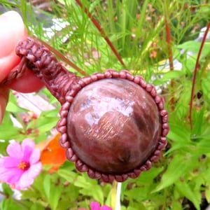 May include: A round, reddish-brown gemstone pendant, intricately woven with a matching cord. The gemstone is encased in a detailed macrame setting. The background features vibrant green foliage and pink flowers.